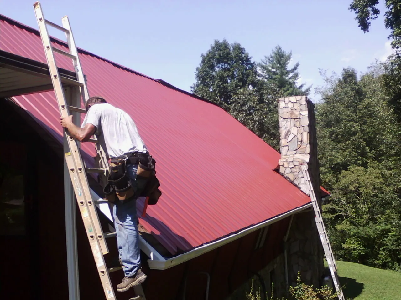 Roofer on a ladder beside a red metal roof and stone chimney — Soffit & Fascia Repair by Roofing Brothers in Nutley
